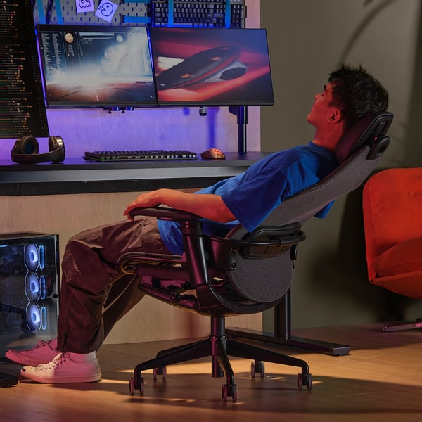 La personne s’allonge dans une chaise de jeu ergonomique avec appui-tête et dossier en maille, face à deux écrans vibrants.
