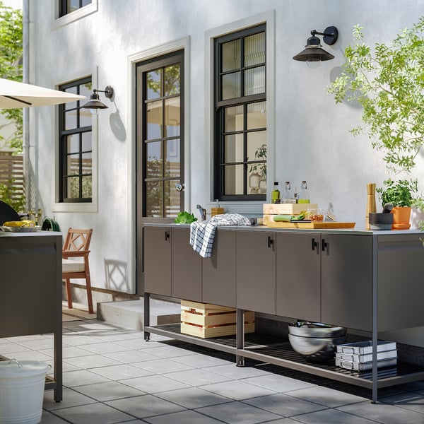 Cuisine extérieure avec unité BÅTSKÄR grise, évier, objets, chaise, plante, grandes fenêtres.
