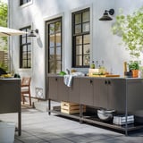 Cuisine extérieure avec unité BÅTSKÄR grise, évier, objets, chaise, plante, grandes fenêtres.