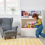 Un enfant jouant avec des jouets sur une bibliothèque basse avec des livres visibles et un tiroir de rangement.
