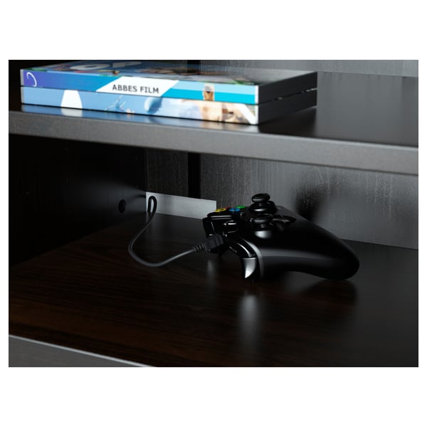 Manette de jeu noire sur meuble TV en bois foncé, montrant les étagères de rangement et la gestion des câbles.