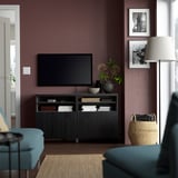 Salon moderne avec banc TV BESTÅ, livres, vases et télévision à écran plat contre mur violet foncé. Mobilier bleu et noir, grand abat-jour.