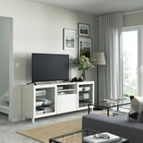 Salon moderne avec banc TV BESTÅ, télévision à écran plat, livres, vase, rideaux gris, lampe et œuvres d’art.