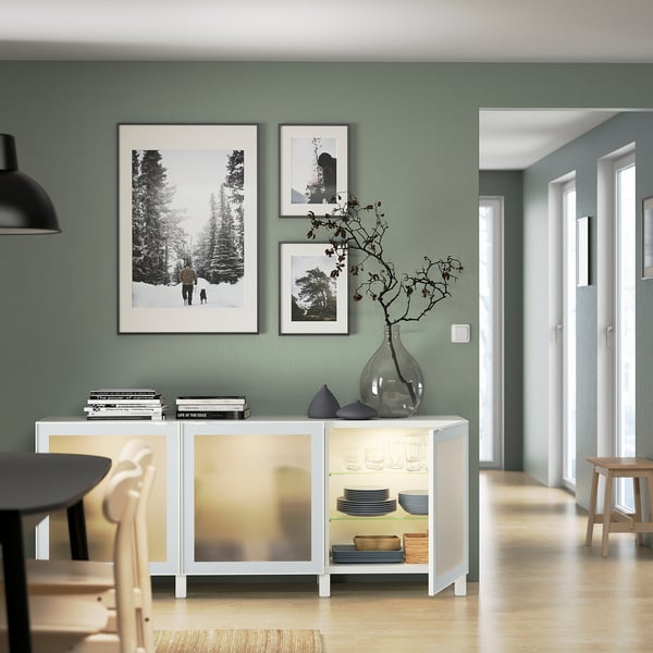 Salle à manger moderne avec buffet BESTÅ, portes vitrées montrant des assiettes, un vase et des photos.