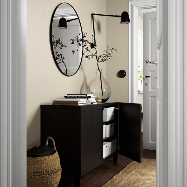 Unité de rangement noire avec portes ouvertes laissant apparaître des étagères et des boîtes. Miroir au-dessus, livres sur le dessus, appliques murales et un vase avec des branches.