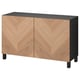 Un buffet ikea BESTÅ en bois clair avec un plateau et des pieds noirs. Il a deux portes présentant un motif en bois de chevron avec des charnières à fermeture douce.