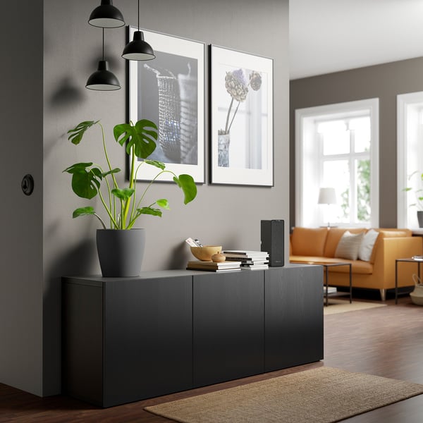 Chambre moderne avec unité de rangement BESTÅ grise, remplie de plantes et de livres. Canapé orange et grandes fenêtres en arrière-plan.