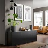 Chambre moderne avec unité de rangement BESTÅ grise, remplie de plantes et de livres. Canapé orange et grandes fenêtres en arrière-plan.