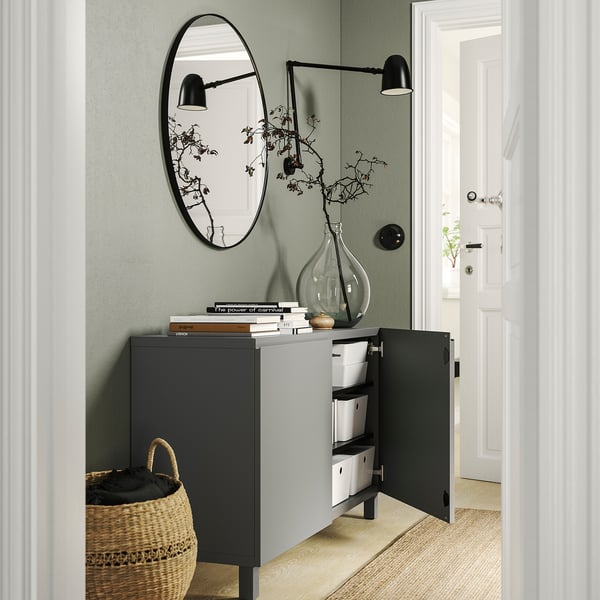 Entrée moderne avec unité de rangement grise BESTÅ, boîtes blanches, miroir rond, appliques noires et un panier tressé.