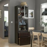 Vitrine en bois sombre avec portes vitrées, montrant des tasses et des verres, positionnée près d’une table à manger.