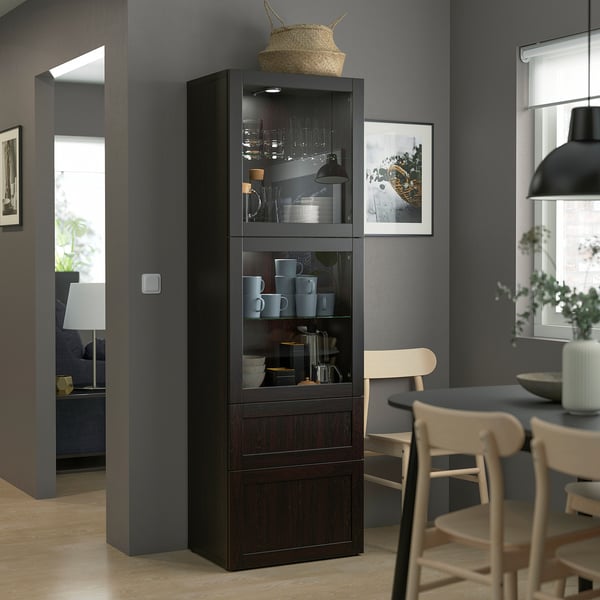 Armoire moderne en bois sombre ikea BESTÅ avec portes en verre, vitrines de tasses et de bols, surmontée d’un panier tressé dans un cadre de salle à manger confortable.