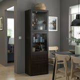 Armoire moderne en bois sombre ikea BESTÅ avec portes en verre, vitrines de tasses et de bols, surmontée d’un panier tressé dans un cadre de salle à manger confortable.