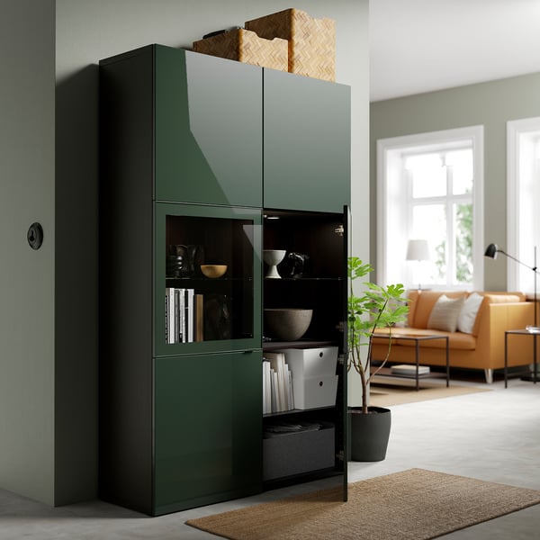 Unité de rangement vert foncé avec portes vitrées exposant des objets, à côté du salon avec canapé et fenêtre.