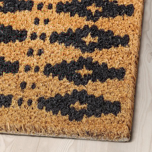 Paillasson à motif géométrique noir sur fibre naturelle, exposé sur un plancher en bois.