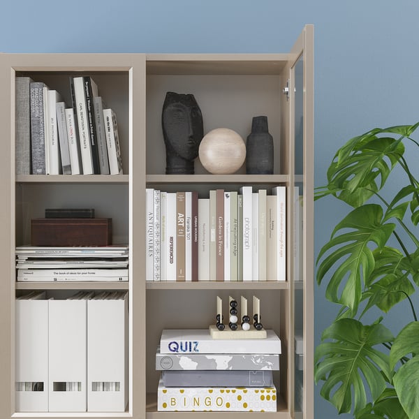 Étagère avec étagères réglables, contenant des livres et des objets de décoration. Des portes vitrées protègent le contenu. Plantez à côté des étagères.