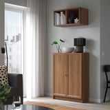 Petite armoire de rangement en bois BILLY près de la fenêtre. Contient des objets décoratifs, sous l’étagère avec une plante en pot.