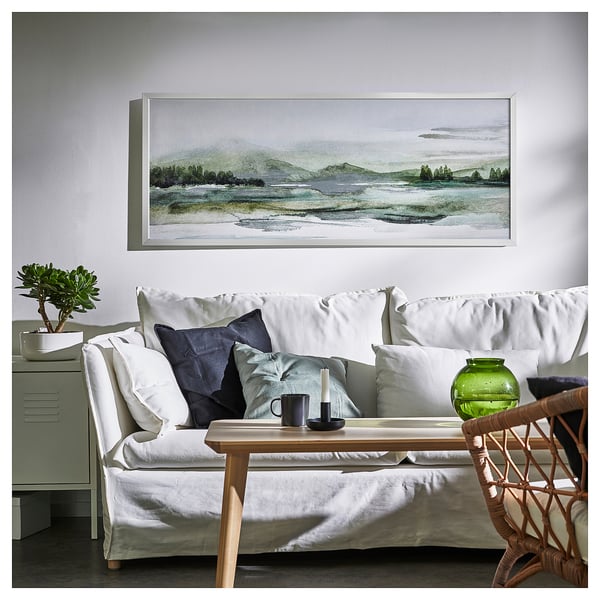 Un grand canapé blanc avec des oreillers noirs et verts se trouve sous une peinture de paysage, avec un vase en verre et un chandelier sur une table basse en bois.