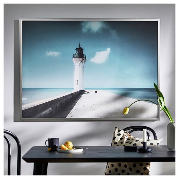 Grand art mural avec phare blanc sur la jetée, ciel bleu. Encadré d’argent, design minimaliste moderne.