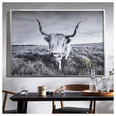 Une grande tête de taureau noir et blanc est suspendue au-dessus d’une table à manger en bois avec 2 chaises, contenant de la verrerie et une bougie.