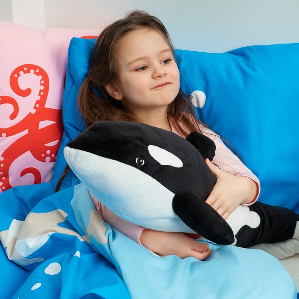 La fille est assise sur un pouf bleu avec un jouet orque en peluche.
