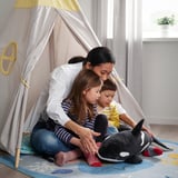 Une femme et deux enfants jouent à l’intérieur d’une tente beige avec une orque en peluche. La tente est de marque BLÅVINGAD.