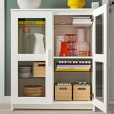 Armoire blanche avec portes vitrées mettant en valeur des vases, des bols, des livres et des boîtes. Les étagères sont réglables et l’armoire est fixée au mur.