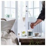 Personne allumant une bougie BUKETTAPEL dans une pièce lumineuse. Il est en verre avec une étiquette verte, sur une table en bois près d’une porte blanche.
