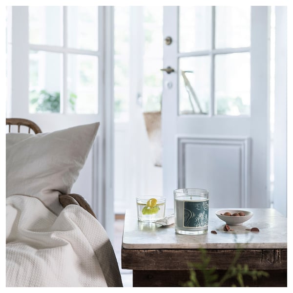 Scène de chambre à coucher confortable avec une bougie verte BUKETTAPEL allumée sur une table rustique, une couverture et un oreiller.