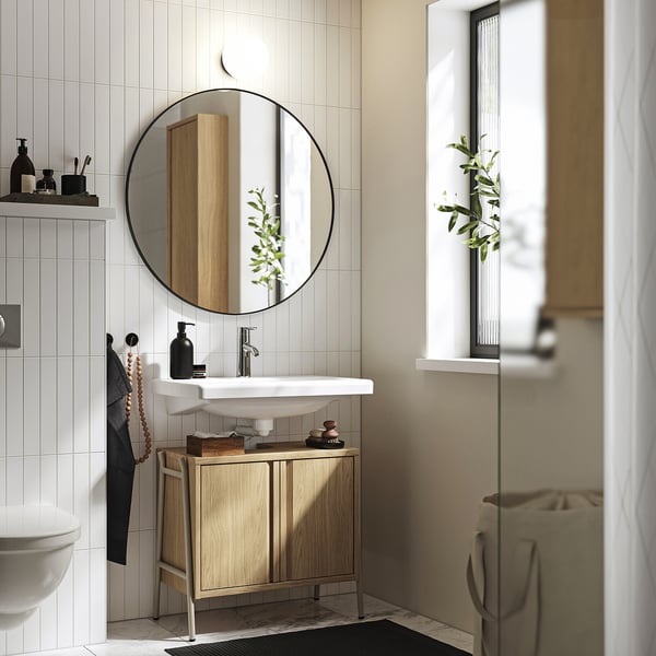 Salle de bain moderne avec armoire en bois sous lavabo. Miroir rond, évier blanc et plante verte. L’armoire s’adapte parfaitement sous l’évier.