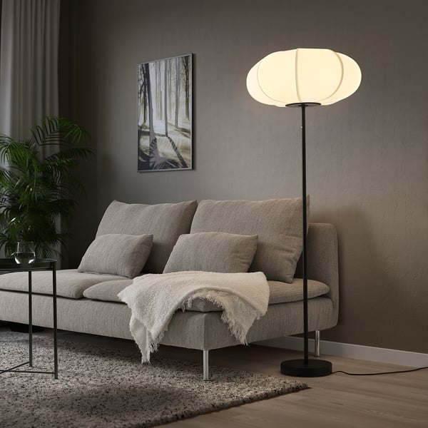 L’élégante lampe BYGGKORN, blanche, à la lumière délicate, se dresse fièrement à côté d’un canapé gris, offrant un éclairage doux et chaleureux à un coin douillet de la pièce.