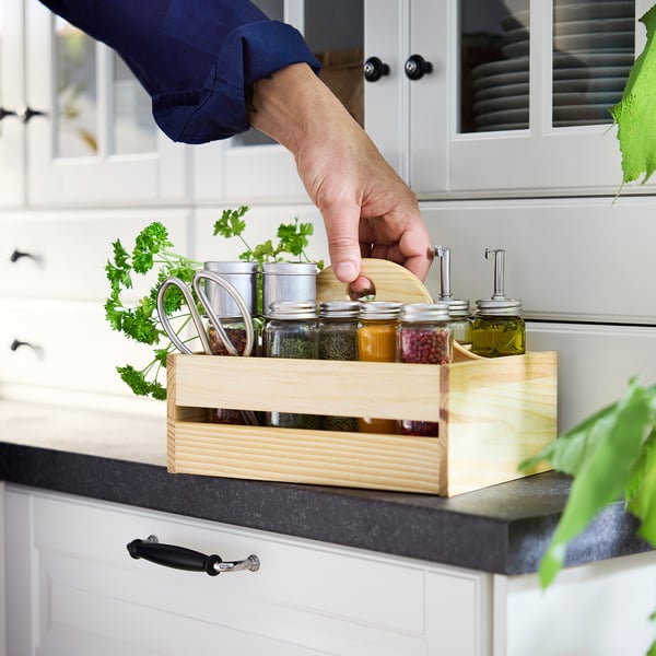 Description de l’image : une main s’étend vers un bocal dans une boîte en bois sur un plan de travail de cuisine, tenant divers bocaux d’épices et des herbes fraîches.