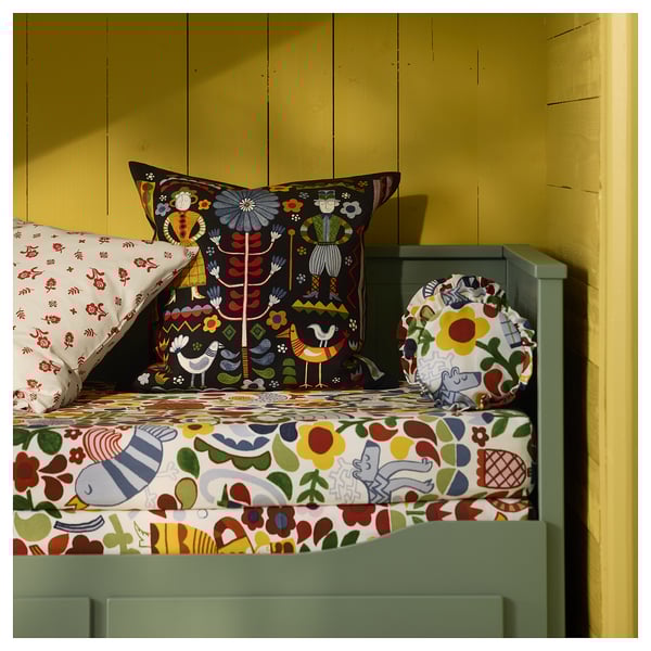 Lit avec des oreillers et une couverture colorés à motifs, avec des motifs d’art populaire, contre un mur jaune.