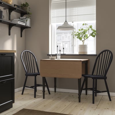 Table à abattants DANDERYD avec chaises noires, compacte pour les petits espaces, plateau en bois clair, pieds en métal noir, plante à côté.