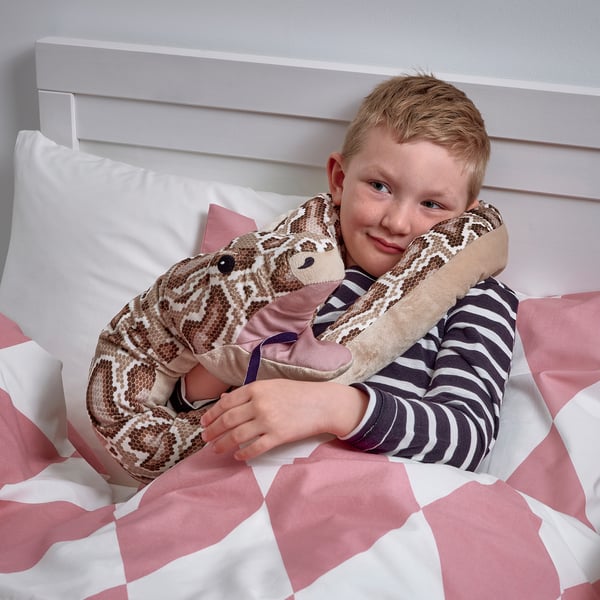 Un garçon serrant dans ses bras un python en peluche dans son lit. Le jouet a une poche dans sa tête pour une main.