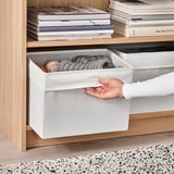 Personne ouvrant une boîte de rangement en tissu blanc de l’étagère, révélant des articles tricotés à l’intérieur. La boîte est étiquetée DRÖNA.