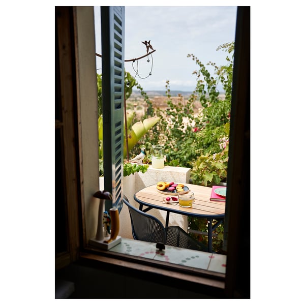 Petite table de balcon et chaises avec des boissons et de la nourriture sur une table, vue de l’intérieur. Volets verts, jardin luxuriant, ciel bleu.