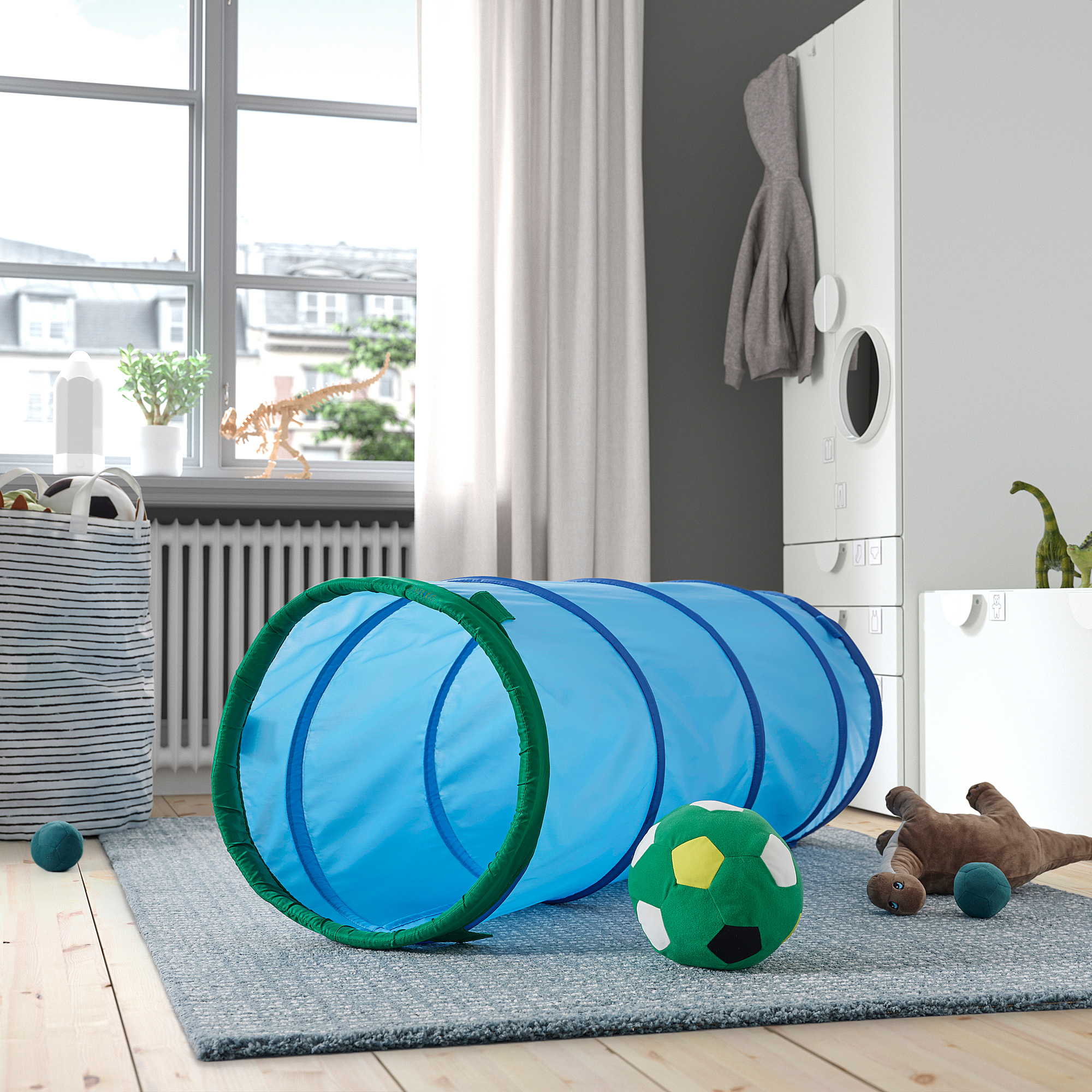 Un tunnel de jeu DVÄRGMÅS bleu pour les enfants, en polyester recyclé, sur un tapis avec des jouets. Il est plus grand que le dinosaure en peluche à proximité.