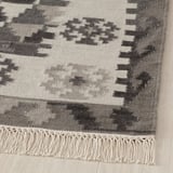 Tapis en laine tissé à la main avec des motifs gris complexes et modernes et des bords frangés sur un plancher de bois franc.