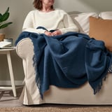 Personne en pull blanc assise sur une chaise beige, recouverte d’un plaid bleu foncé à franges de la série DYTÅG.