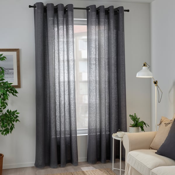 Rideaux en lin gris avec motif d’œillets par fenêtre, filtrant la lumière. A proximité, une lampe blanche et une chaise beige avec des oreillers.