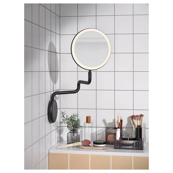 Miroir noir moderne avec lumières intégrées, monté sur un mur, entouré d’accessoires de maquillage sur un comptoir de salle de bain carrelé de blanc.