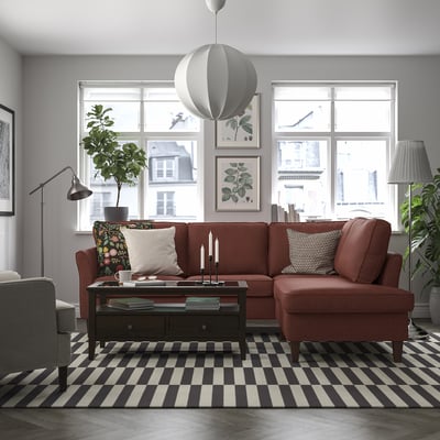 Salon moderne avec grand canapé rouge rouille EKHOLMA, tapis rayé, suspension grise, plante et vue sur la ville à travers de hautes fenêtres.