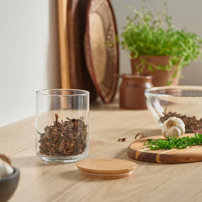 Petit pot EKLATANT avec couvercle en bambou, rempli de champignons séchés. Sur une table de cuisine près des herbes et de la planche à découper.