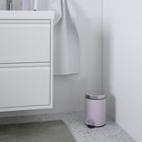 Une salle de bains moderne avec une petite poubelle EKOLN violet clair avec un couvercle argenté positionné entre une armoire blanche et un mur carrelé gris clair.