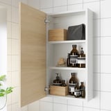 Armoire de salle de bain ouverte exposant des bouteilles, des boîtes en bois et un chiffon gris, avec porte en chêne et deux étagères.