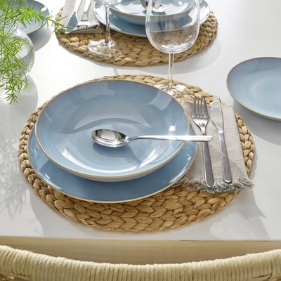 Une table avec des assiettes et des bols bleu clair sur des sets de table tissés, y compris des couverts et des verres à vin transparents.