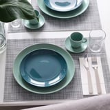 Une table dressée avec des assiettes bleu-vert, des tasses, des verres et des ustensiles métalliques sur des sets de table tissés, prête à être dégustée.