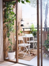 Petit balcon confortable avec table et chaises en métal blanc, plantes vertes et tapis naturel, vu de l’intérieur.