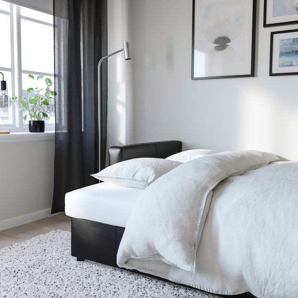 Lit avec des draps blancs, une tête de lit grise, des rideaux noirs et une plante en pot près d’une fenêtre.