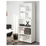 Unité de rangement de bureau blanche avec plusieurs étagères, contenant des classeurs et des boîtes, positionnée à côté d’un bureau.